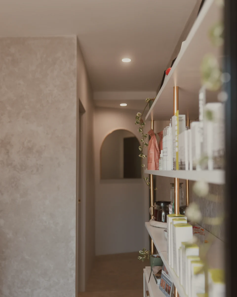 A bright hallway with a limewashed wall, an arched doorway and softly blurred shelves of skincare