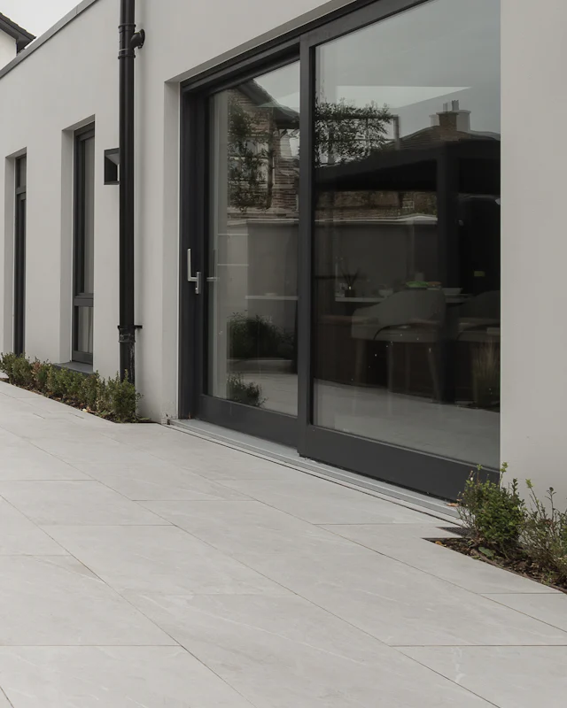 The garden side of a house in smooth pale render, with a tall black-framed sliding door onto a terrace of large grey tiles