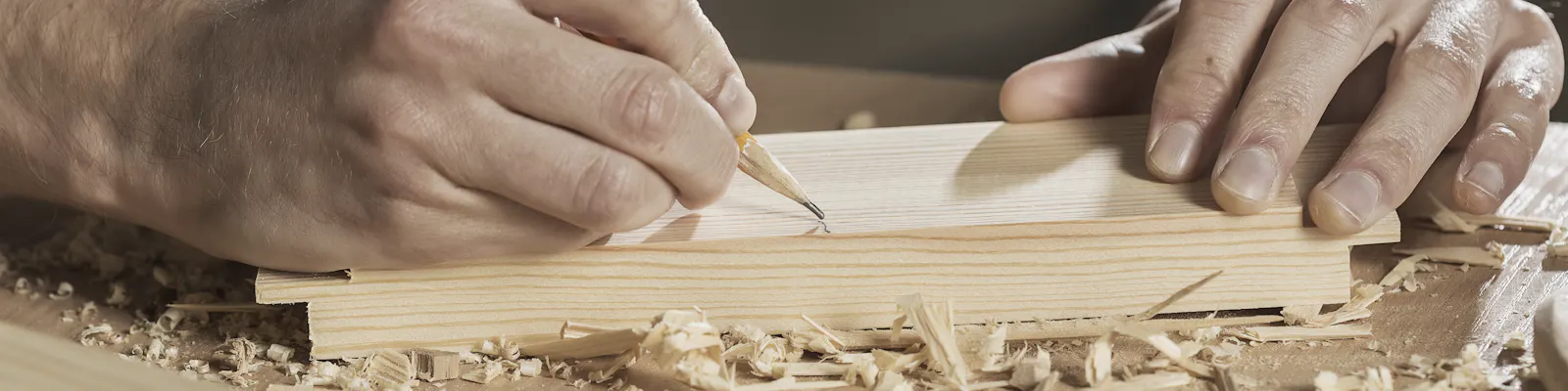 Two hands marking a line in pencil on a pine board, wood shavings on the bench