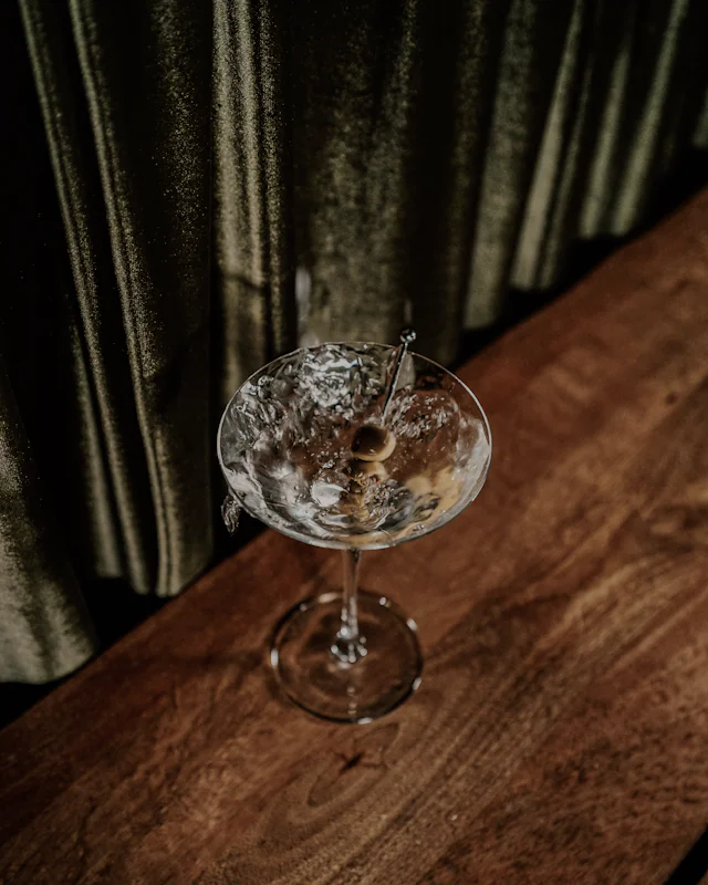 A martini with olives on a dark wooden bar, a green velvet curtain behind it