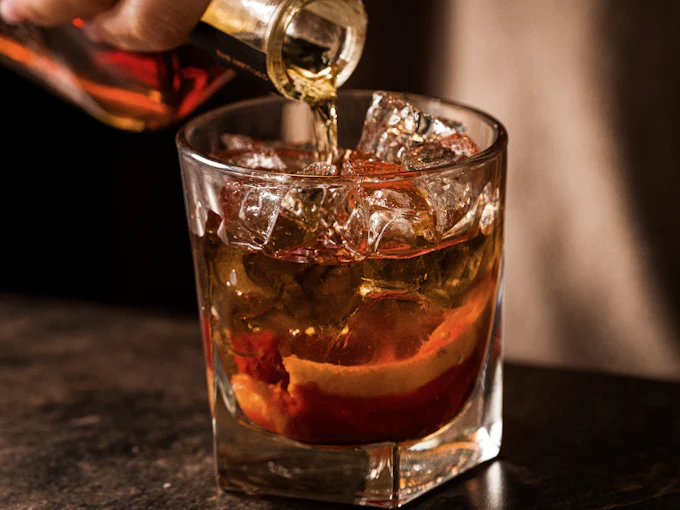 A hand pouring an amber spirit over ice and orange peel in a tumbler, on a dark counter
