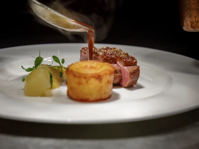 A spoon pouring dark sauce over a main course: sliced meat with a crisp crust, a fondant potato and a poached pear