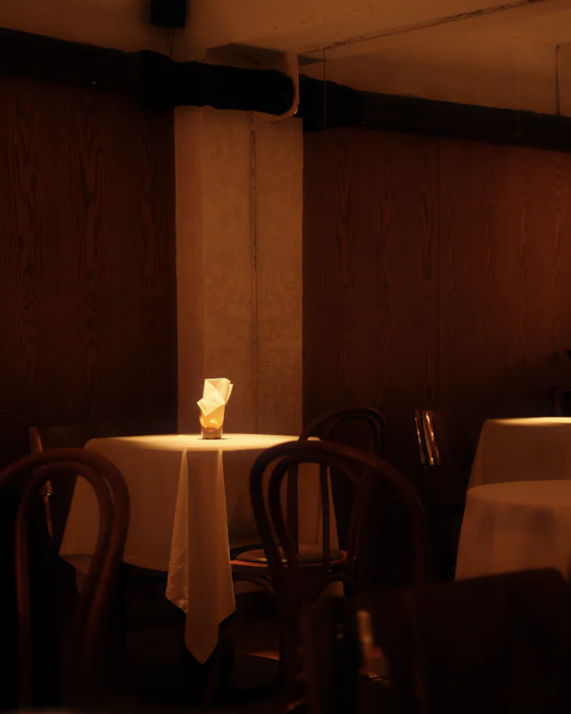 A single candle on a table with a long white cloth in a dim, wood-panelled room, bentwood chairs around it