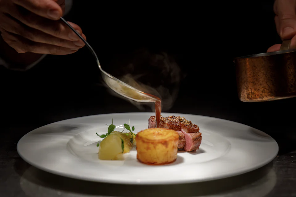 A cook’s hands spooning sauce over a plated main course at a steel pass, a copper pan beside it