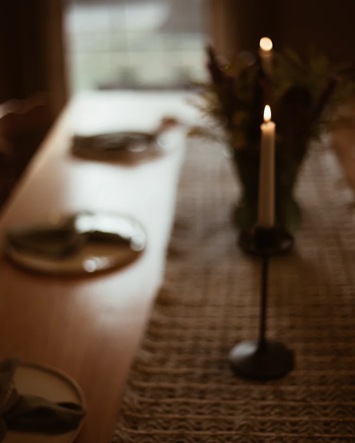 A long oak table in soft focus: lit taper candles, a vase of dried flowers and plates along a woven runner