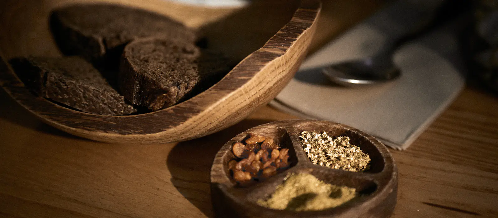 Slices of dark bread in a wooden bowl beside a small dish of seeds and spices