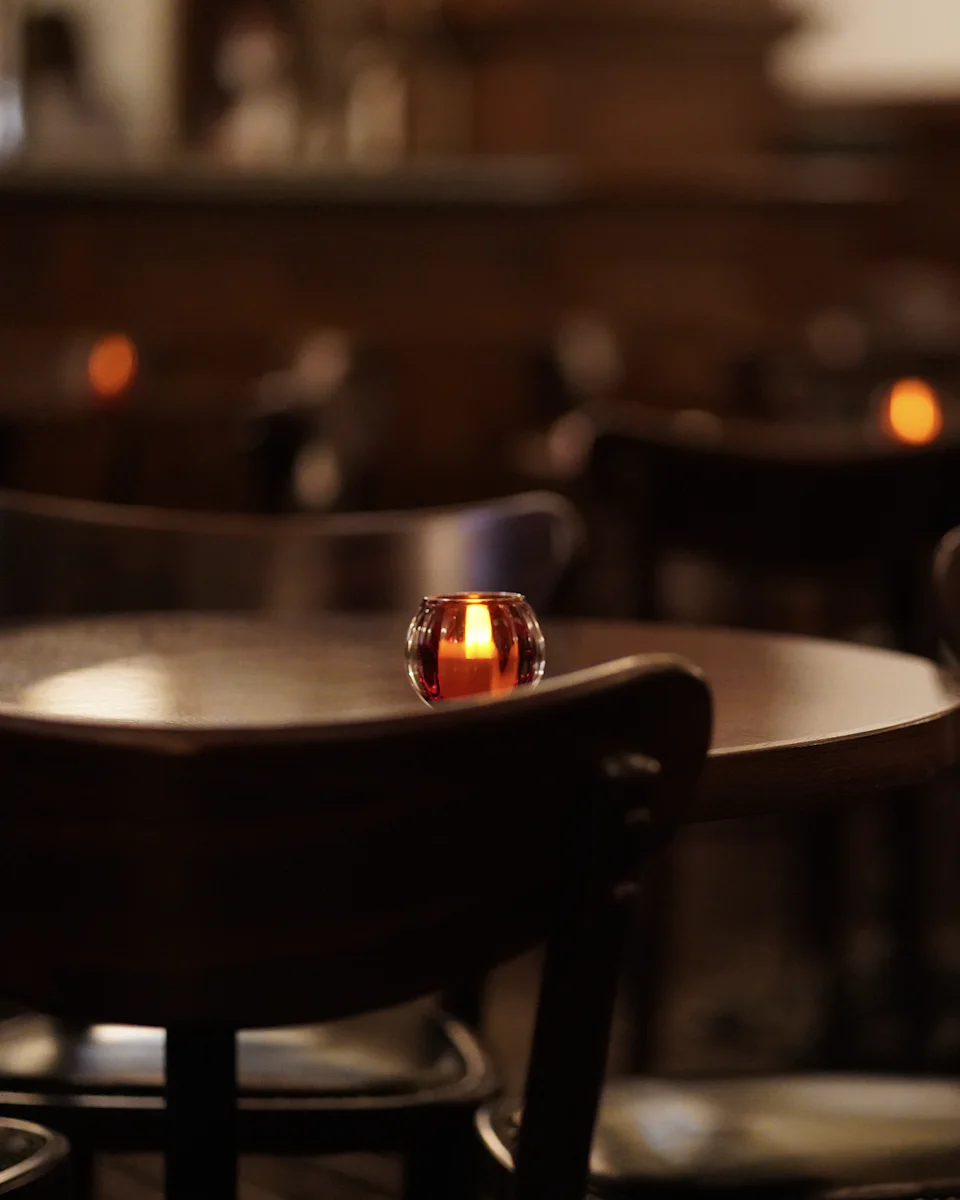 A small round table with a tealight in a red glass and dark bentwood chairs, more candlelit tables blurred behind it