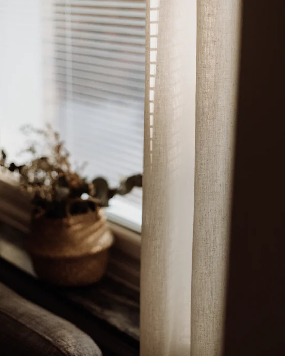 A beige linen curtain beside a window with blinds, a basket of dried flowers on the sill
