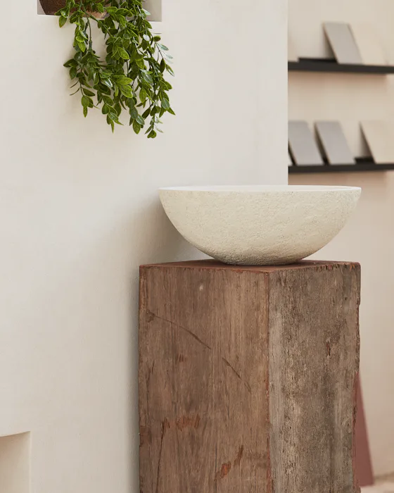 A pale stone bowl on a weathered timber plinth against a limewashed wall, a trailing plant above