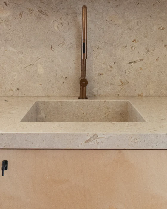 A square washbasin cut into a pale stone counter, a brass tap against the stone wall behind it and light wooden cabinet doors below