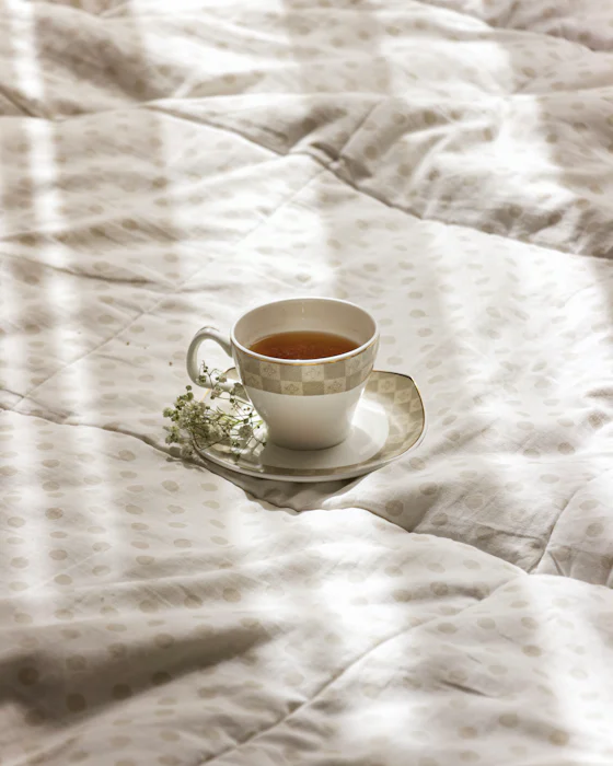A cup of tea on a saucer with a sprig of small white flowers, on rumpled linen in striped window light