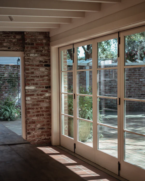 Sunlight through tall folding glass doors onto a tiled floor, beside an old brick wall and under white-painted timber beams