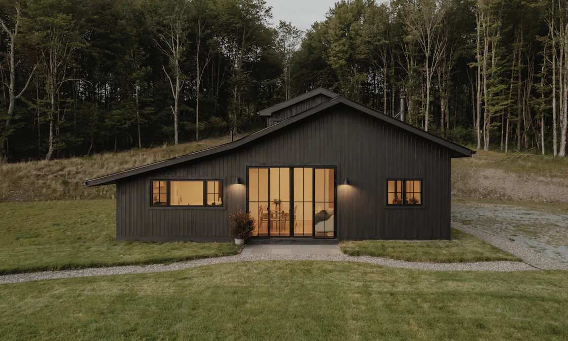 A single-storey house clad in black vertical boards, lit from inside at dusk, with a wide glazed door onto a lawn