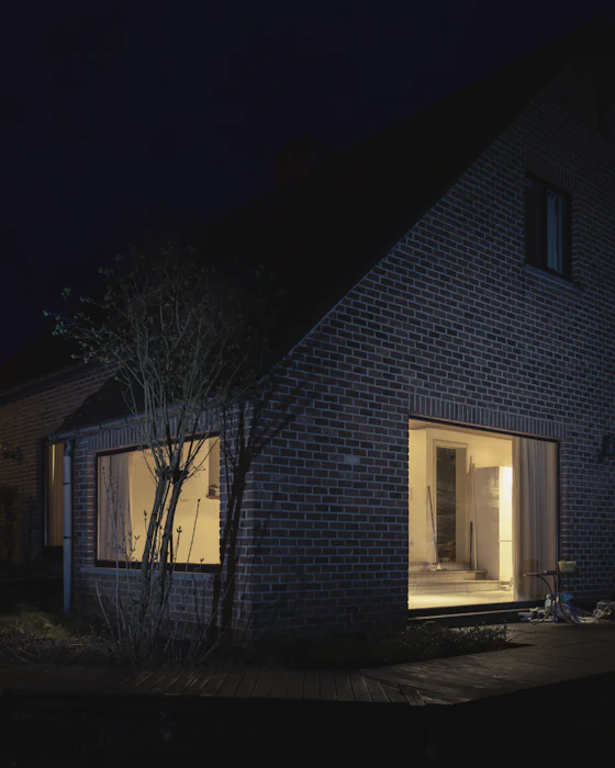 A grey-brick house at night, light from two wide window openings falling onto a wooden deck
