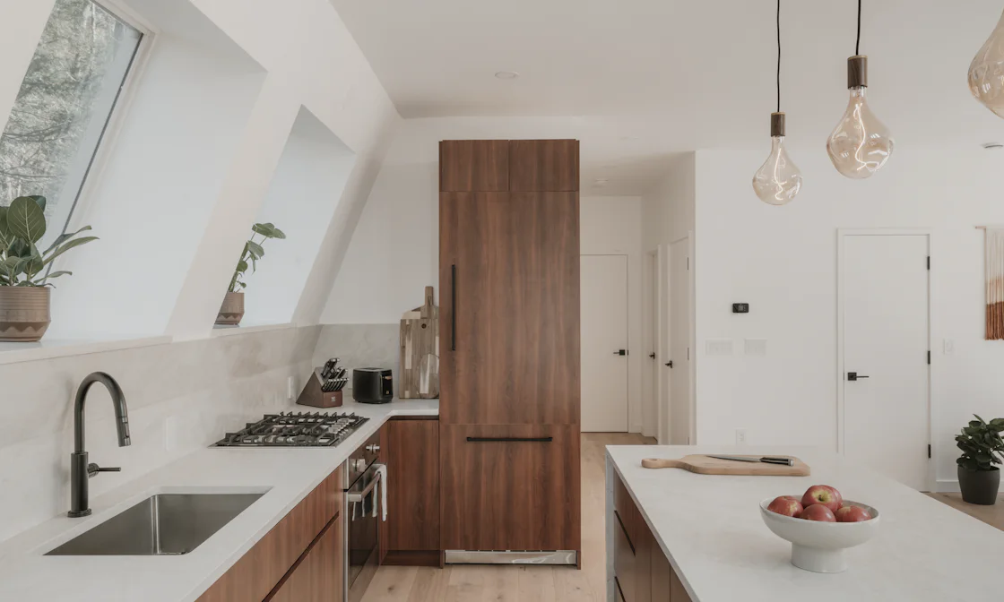 A white kitchen under a sloping ceiling with roof windows, a sink below them and a tall wood-fronted cupboard
