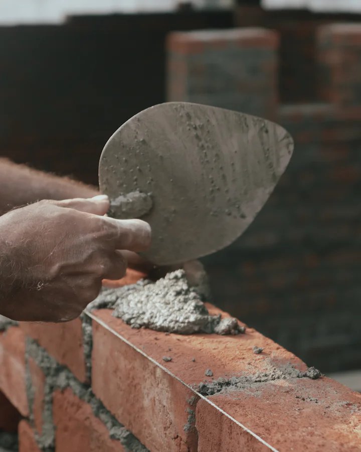 A hand spreading mortar with a trowel on a course of red bricks