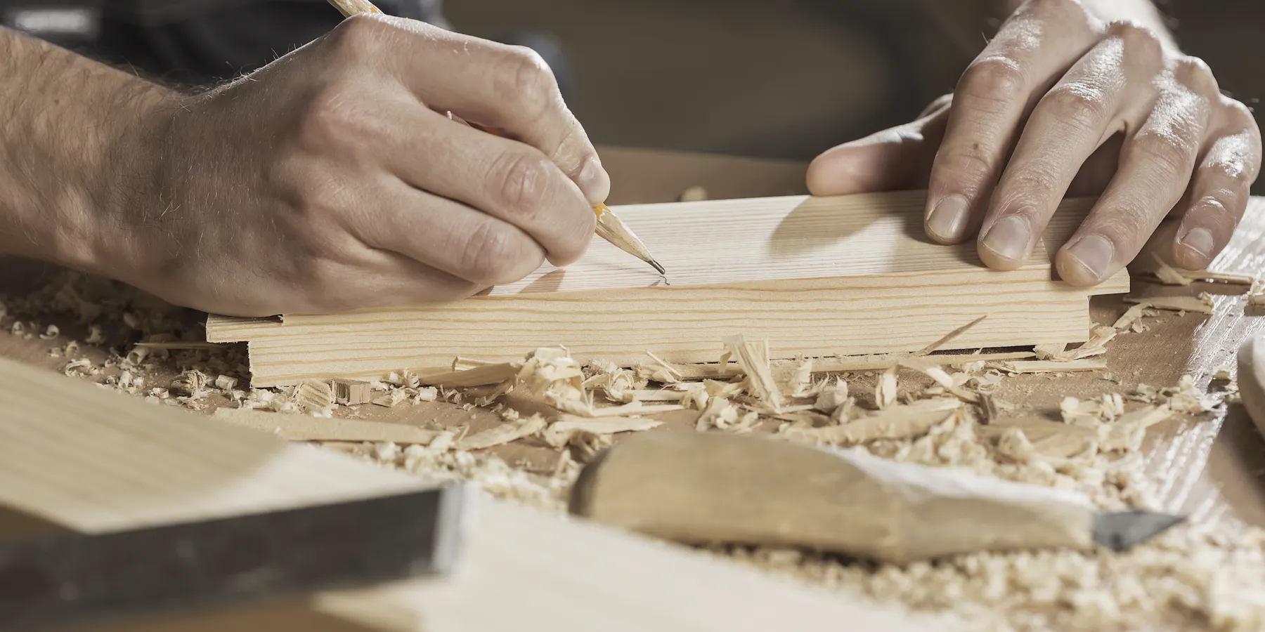 Two hands marking a line with a pencil on a pine board, wood shavings around them