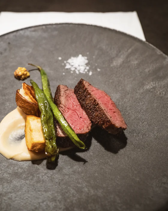 Slices of pink meat with a roast shallot, green peppers and a purée on a dark plate