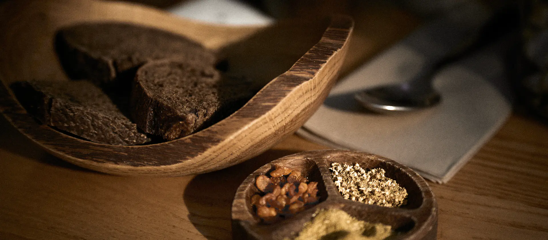 Slices of dark bread in a wooden bowl beside a small dish of seeds and spices