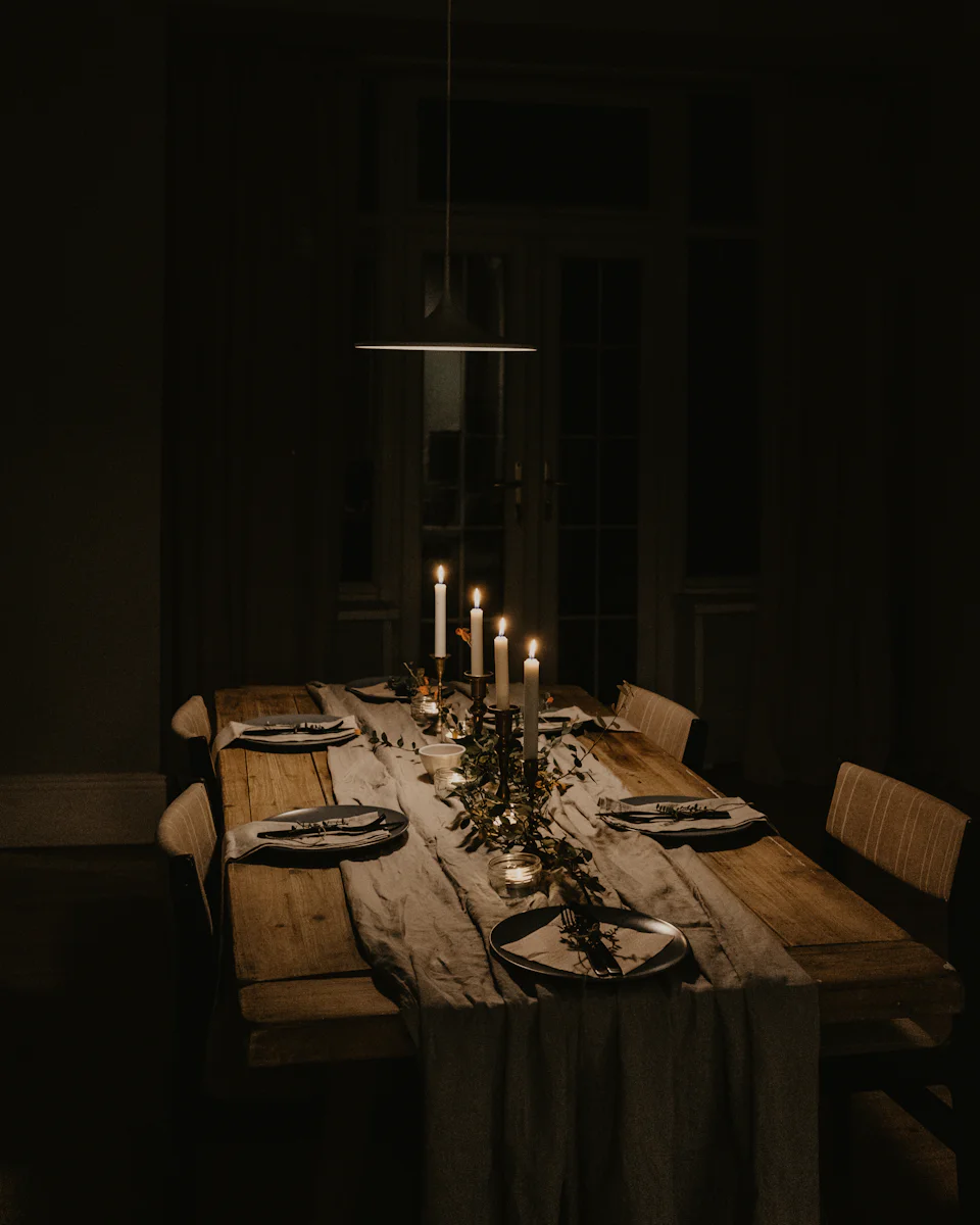 One long wooden table set for eight in a dark room with curtains, tall candles and greenery on a linen runner, a pendant lamp above