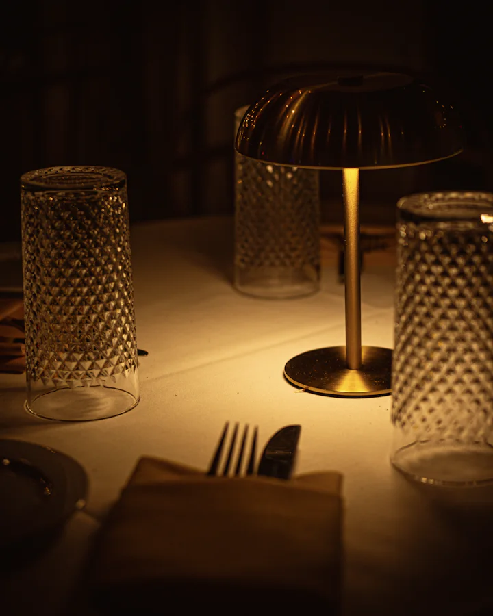 A table laid for the evening: a small brass lamp lit on a white cloth, crystal tumblers and a fork and knife in a folded napkin